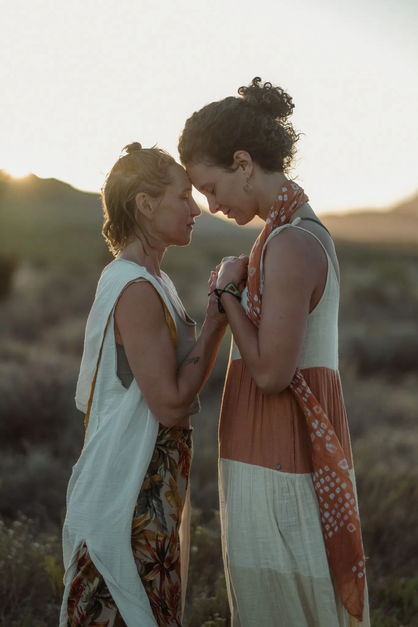 Two women standing forehead to forehead at sunset, hands clasped